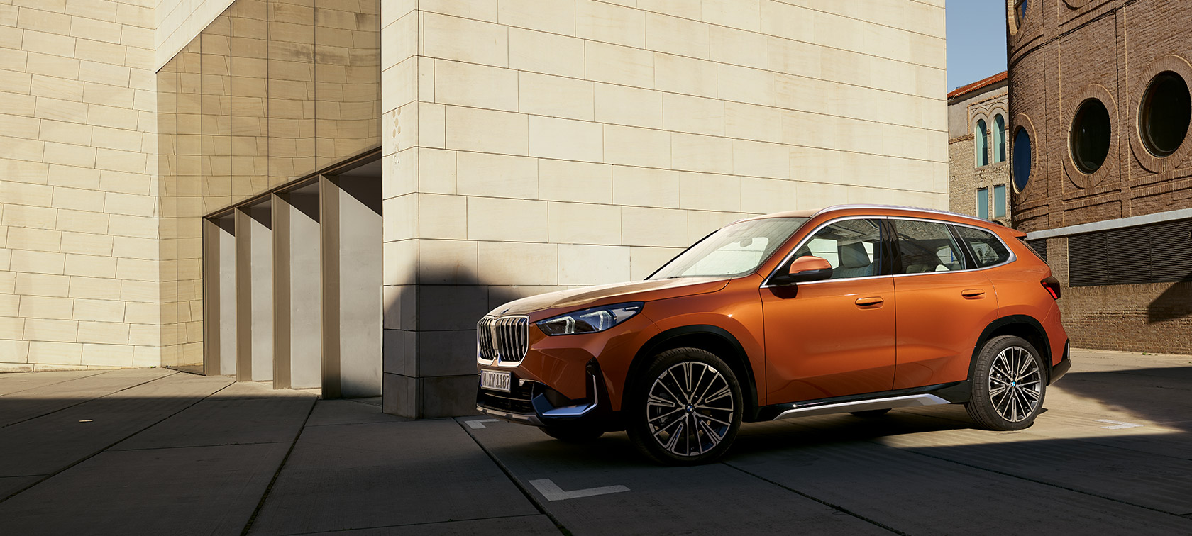 BMW U11 X1 xLine в цвете Utah orange metallic, вид спереди с поворотом в три четверти напротив здания